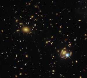A cluster of various types of galaxies, visible as oval shapes in clumps. Two spiral galaxies are especially prominent near the center, and another on the right, where the lensing effect is best visible as a reddish curve with a bright star next to it and the same distorted galaxy appearing several times.
